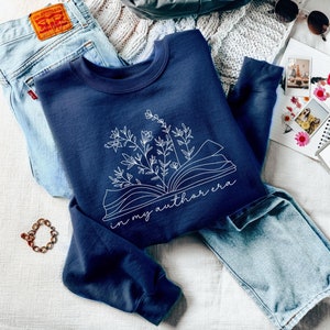 May include: A navy blue sweatshirt with a white outline of an open book with flowers growing from it. The text "in my author era" is printed below the book.