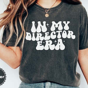 May include: A dark gray t-shirt with the text "IN MY DIRECTOR ERA" in white, retro-style lettering.