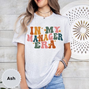 May include: A light grey t-shirt with the text "In My Manager Era" in colorful retro-style lettering with stars. The shirt is being worn by a person.