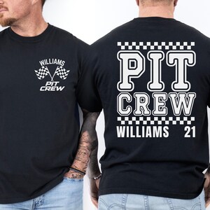 Op de afbeelding: Zwart T-shirt met witte tekst en geruit ontwerp. De tekst luidt "PIT CREW WILLIAMS 21" op de achterkant en "WILLIAMS PIT CREW" op de voorkant met een geruite vlaggrafiek.