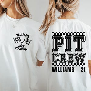 Op de afbeelding: Wit T-shirt met zwarte tekst en een geruit patroon. De tekst luidt "PIT CREW WILLIAMS 21" en "WILLIAMS PIT CREW" met een geruite vlaggrafiek.