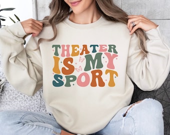 May include: A cream-colored sweatshirt with the text "THEATER IS MY SPORT" in colorful, retro-style lettering. The letters are outlined in black and have a slightly raised, 3D effect. There are small pink butterflies scattered around the text.