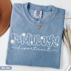 May include: A blue jean coloured t-shirt with the words "Radiology department" in white, decorated with small daisy flowers. A gold necklace with a pendant rests on the shirt. The shirt is a Comfort Colors brand.