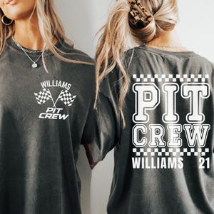 Op de afbeelding: Twee grijze T-shirts met witte tekst en afbeeldingen. Het voorste shirt zegt "Williams Pit Crew" met een geruite vlag afbeelding. Het achterste shirt zegt "Pit Crew Williams 21" met een geruite rand.