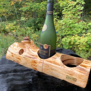 Peut inclure: Un porte-bouteille de vin en bois rustique avec deux ouvertures, l'une contenant une bouteille de vin verte avec un bouchon. Le support est en bois clair et porte une petite plaque en laiton avec l'inscription "BUCCI".