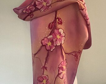 May include: A rectangular silk scarf featuring a hand-painted floral design with pink blossoms on thin brown branches. The background transitions from a deep plum purple at the top to a soft peach and light pink gradient toward the bottom.