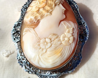 May include: Oval cameo brooch featuring a carved profile of a woman with flowing hair and floral accents. The cameo is set in a silver-toned filigree frame. The woman's profile is in shades of cream and peach.