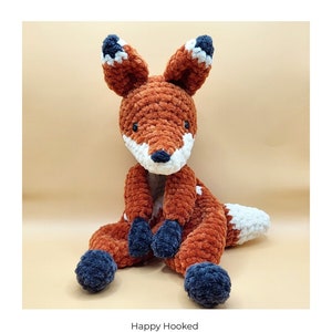 May include: A crocheted fox amigurumi toy with orange, white, and blue yarn. The toy is sitting on a light brown surface. The text "Happy Hooked Crochet pattern / Haakpatroon Frankie Fox / Frankie Vos English + Nederlands PDF" is below the toy.