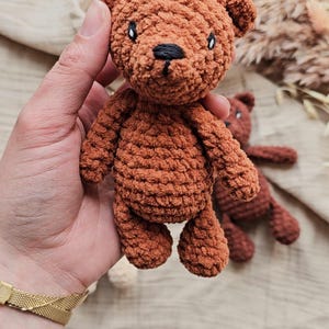 May include: A handmade brown crochet teddy bear with black eyes and nose. The bear is held in a hand, with another bear visible in the background. The bear is a soft toy.