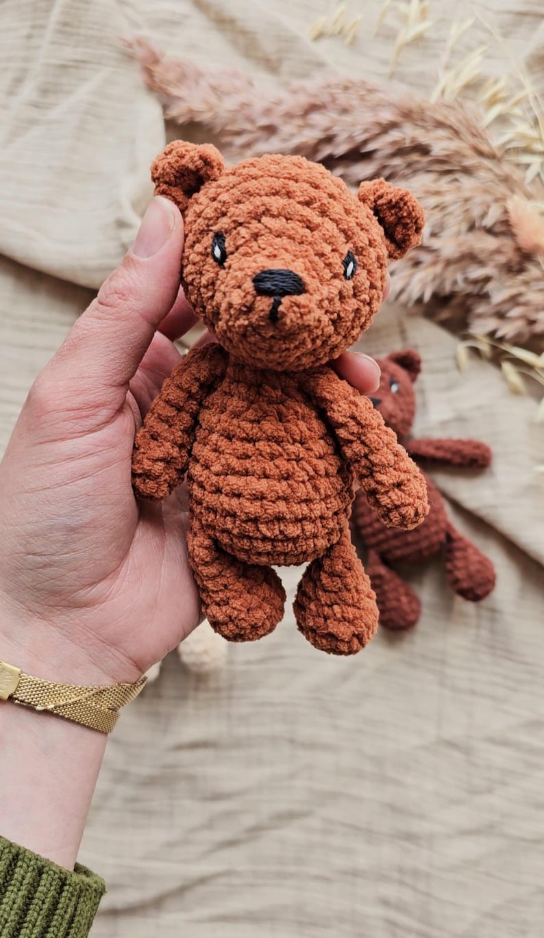 May include: A handmade brown crochet teddy bear with black eyes and nose. The bear is held in a hand, with another bear visible in the background. The bear is a soft toy.