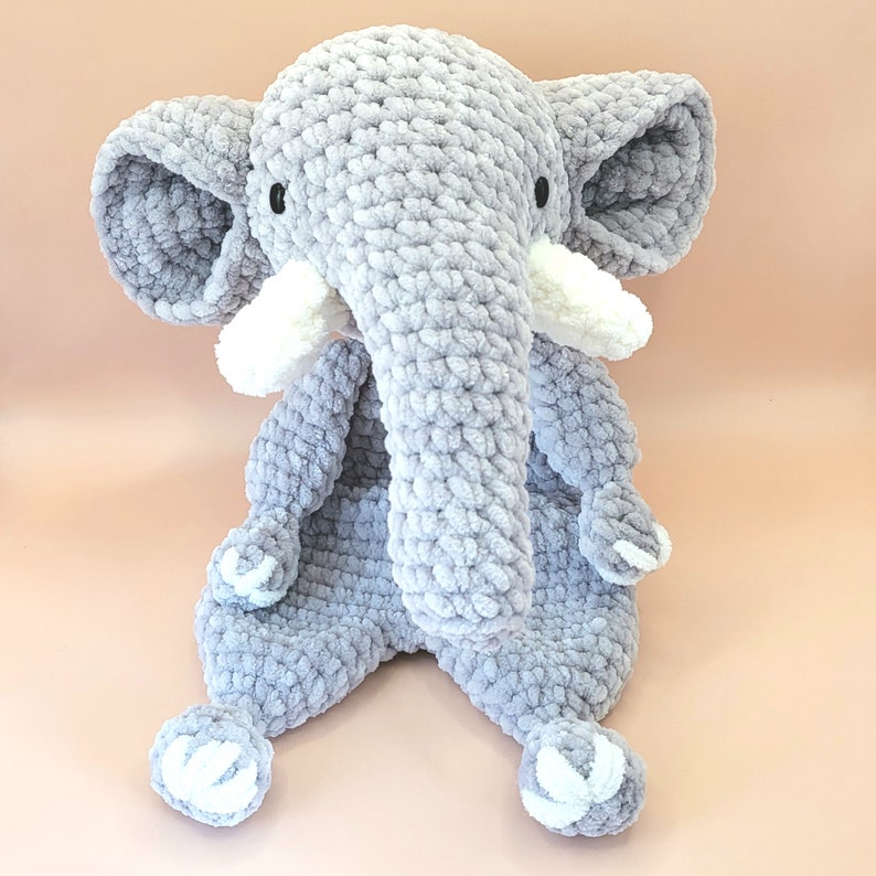May include: A gray crocheted elephant stuffed animal with white crocheted ears and feet. The elephant has a long trunk and is sitting on a light pink surface.