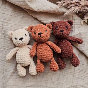 May include: Three handmade crochet teddy bears in cream, orange, and brown. Each bear has a black nose and eyes, with a textured, plush appearance. The bears are arranged on a neutral-toned fabric background.