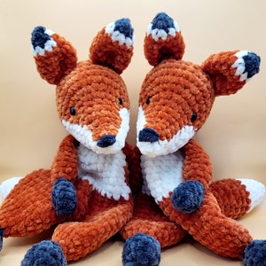 May include: Two crocheted fox plushies with orange and white fur. The foxes are sitting side by side and have black eyes and noses.