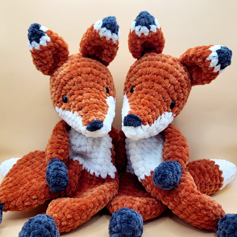 May include: Two crocheted fox plushies with orange and white fur. The foxes are sitting side by side and have black eyes and noses.