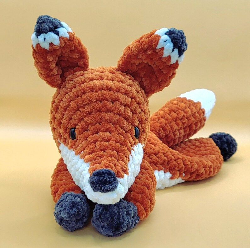 May include: A crocheted fox plush toy with orange and white fur, black eyes, and a black nose. The toy is lying on its side with its head resting on its paws.