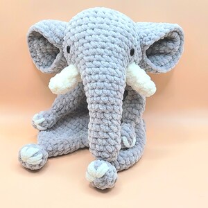 May include: A gray crocheted elephant plush toy with white accents. The elephant is sitting with its legs crossed and its trunk curled up.