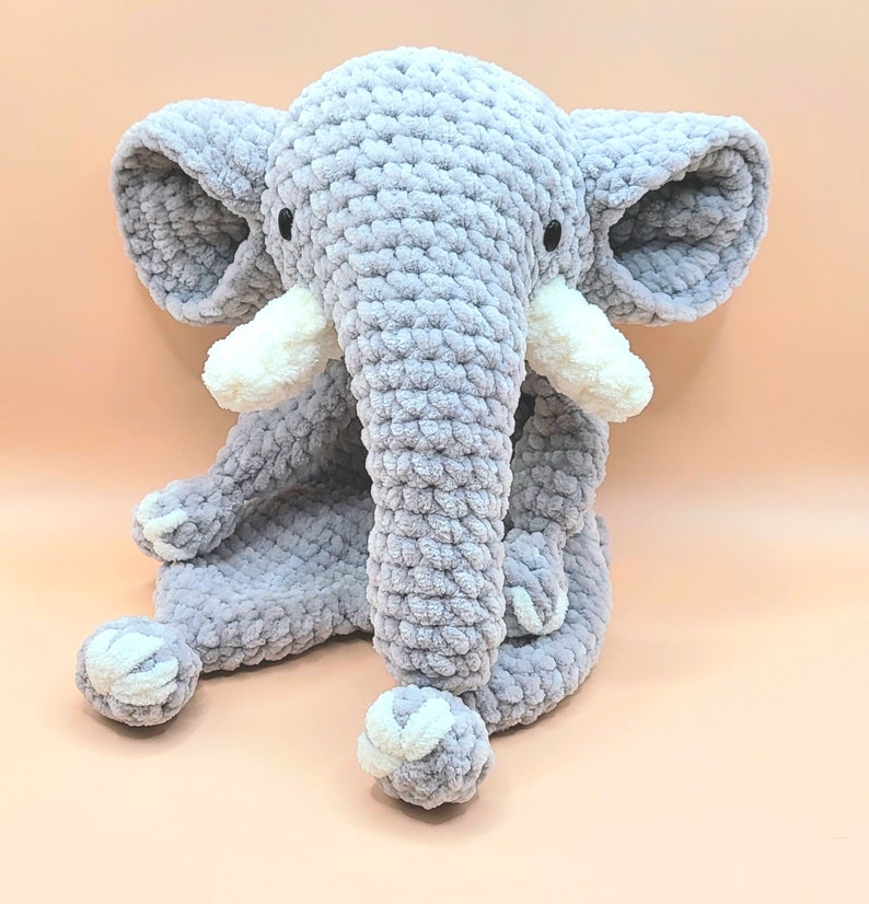 May include: A gray crocheted elephant plush toy with white accents. The elephant is sitting with its legs crossed and its trunk curled up.