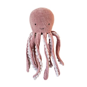 Pode incluir: Um brinquedo de pelúcia de polvo de crochê rosa com detalhes brancos nos tentáculos. O polvo tem uma cara sorridente.