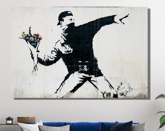 May include: A black and white stencil art print of a person throwing a bouquet of flowers. The person is wearing a black mask and a black cap. The background is a white wall with some visible texture.
