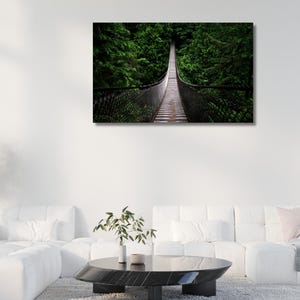 May include: A framed canvas print of a suspension bridge in a forest. The bridge is made of wood and metal, and it is surrounded by lush green trees. The artwork is displayed on a white wall above a white sofa.
