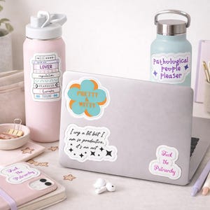 May include: A light pink water bottle with a white lid and a sticker that says "LOVER". A light blue water bottle with a silver lid and a sticker that says "Pathological People Pleaser". A laptop with stickers that say "Pretty & Witty", "I cry a lot but I am so productive, it's an art", and "Fuck The Patriarchy".