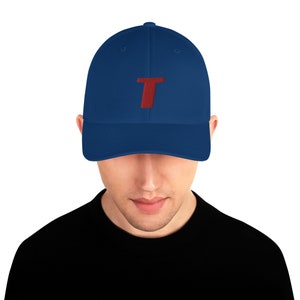 Puede incluir: Una gorra de béisbol azul marino con una letra "T" bordada en rojo en la parte delantera.