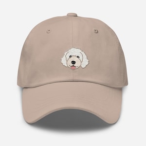 Puede incluir: Una gorra de béisbol beige con un rostro de perro blanco bordado con una lengua rosa.