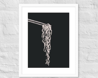 May include: A black and white photograph of a single strand of noodles being held up by chopsticks against a black background.