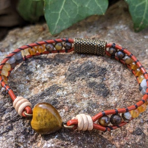 May include: A leather bracelet with a heart-shaped tiger's eye stone charm. The bracelet is made with red and black leather cord and features various gemstone beads in shades of brown, orange, and white.