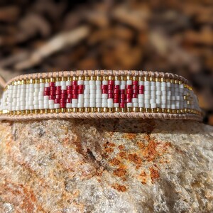 May include: A white beaded bracelet with two red heart designs. The bracelet is made with a brown leather strap.