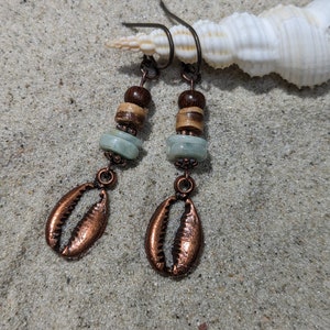 Op de afbeelding: Koperen schelp oorbellen met turquoise en houten kralen. De oorbellen liggen op een zanderige achtergrond met een schelp.