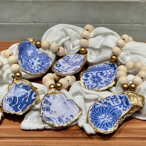 May include: A collection of handcrafted napkin rings. Each ring features a blue and white porcelain shard with a gold-painted edge, strung on a strand of wooden beads. The porcelain pieces display various floral and patterned designs.