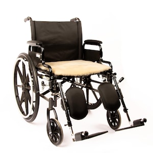 May include: A black manual wheelchair featuring a black frame, black seat and backrest, and a beige sheepskin seat cover. The wheelchair includes two large rear wheels, two smaller front swivel casters, armrests, and adjustable leg rests.