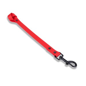 May include: A bright red strap featuring a knotted end and a black metal swivel lobster clasp. The strap is secured with two black rivets along its length, providing a functional connection point for various items in a simple, linear design.