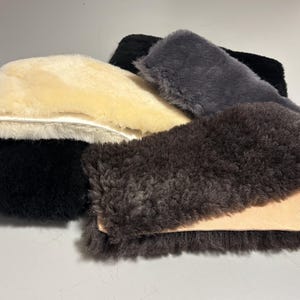 Könnte beinhalten: Eine Sammlung von Schaffell-Pads in verschiedenen Farben, darunter Creme, Schwarz, Grau und Braun. Die Pads haben eine weiche, flauschige Textur und werden wahrscheinlich für Komfort oder Schutz verwendet. Das Bild zeigt die natürlichen Farb- und Texturvariationen.