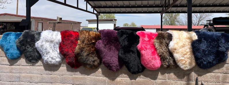 Pu&ograve; includere: Una fila di 12 coprisedia in pelliccia sintetica di vari colori, tra cui blu, grigio, bianco, rosso, marrone, viola, nero, rosa, beige e blu navy. Le coprisedia sono appese a un muro di mattoni.