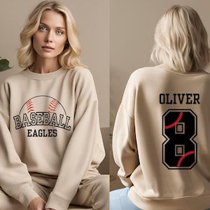 May include: A beige sweatshirt with a baseball graphic and the text "Baseball Eagles" on the front. The back of the sweatshirt has the name "Oliver" and the number "8" with a baseball graphic inside.