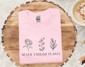 May include: A light pink sweatshirt with the text "NEVER ENOUGH PLANTS" and three black plant illustrations. The sweatshirt is folded on a wooden tray, with a cup of coffee and a cream-colored blanket in the background.