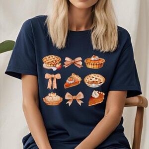 May include: A navy blue t-shirt with a graphic design of various pies and bows. The pies are decorated with fruit and whipped cream.