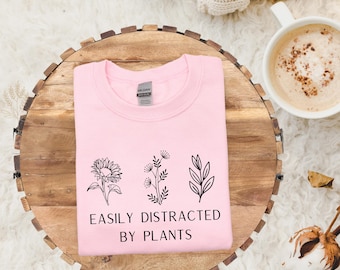 May include: A light pink sweatshirt with the text "EASILY DISTRACTED BY PLANTS" and three black plant illustrations. The sweatshirt is folded on a wooden tray, with a cup of coffee and a cream-colored blanket in the background.