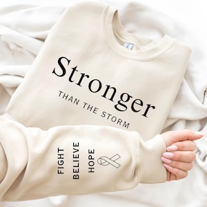 May include: A beige sweatshirt with the text "Stronger than the storm" printed on the front. The left sleeve has the words "Fight, Believe, Hope" and a ribbon printed on it.