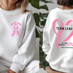 Peut inclure: Sweat-shirt blanc avec des graphiques de ruban rose. Le devant pr&eacute;sente "Support Squad" et "Team Leah" avec un motif de ruban. Le dos affiche un grand c&oelig;ur de ruban rose avec "Team Leah" et le texte "In this family, nobody fights alone." #breastcancerawareness