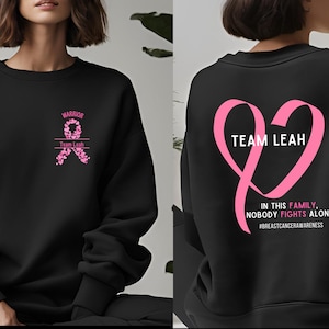 Peut inclure: Sweat-shirt noir avec un motif de ruban rose et du texte. L'avant pr&eacute;sente un ruban avec le mot "Warrior" et "Team Leah". Le dos a un ruban en forme de c&oelig;ur rose avec le texte "Team Leah" et "In this family, nobody fights alone".