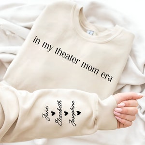 May include: A beige sweatshirt with the text "in my theater mom era" printed on the front and the names "Jane", "Elizabeth", and "Josephine" printed on the sleeve with a heart symbol next to each name.