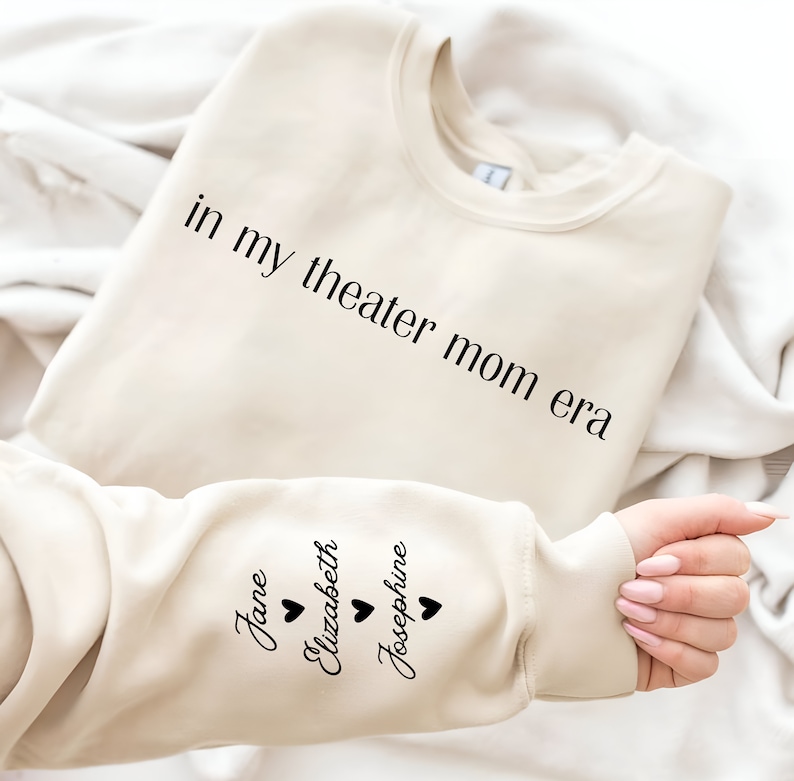 May include: A beige sweatshirt with the text "in my theater mom era" printed on the front and the names "Jane", "Elizabeth", and "Josephine" printed on the sleeve with a heart symbol next to each name.