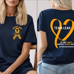 May include: Navy blue t-shirt with the text "SUPPORT SQUAD" and "Team Leah" with a yellow ribbon design. The back of the shirt features a large yellow heart with the words "TEAM LEAH" and the phrase "IN THIS FAMILY, NOBODY FIGHTS ALONE".