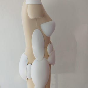 May include: A beige dress form with a black neck and stand, featuring white, curved, padded shapes attached to the form. The shapes are various sizes and are arranged to mimic the contours of a body. The dress form is positioned against a white wall.