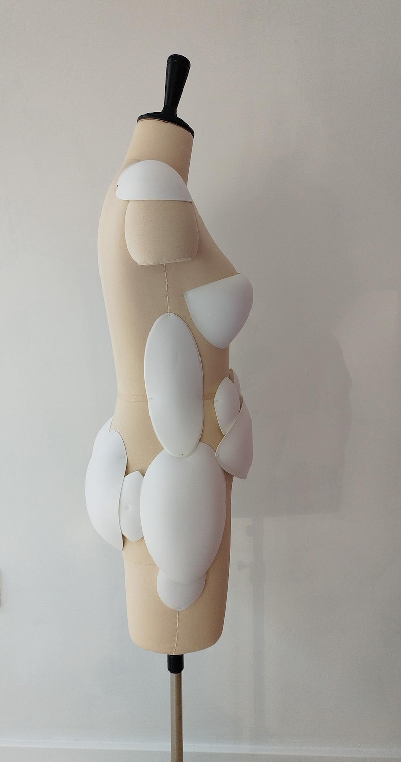 May include: A beige dress form with a black neck and stand, featuring white, curved, padded shapes attached to the form. The shapes are various sizes and are arranged to mimic the contours of a body. The dress form is positioned against a white wall.