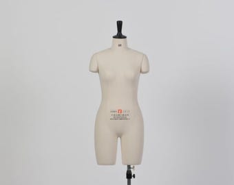 May include: A beige dress form with a black neck and stand. The form has a torso shape with short sleeves and a partial leg form. The form has a label with text on the front. The dress form is on a black stand.