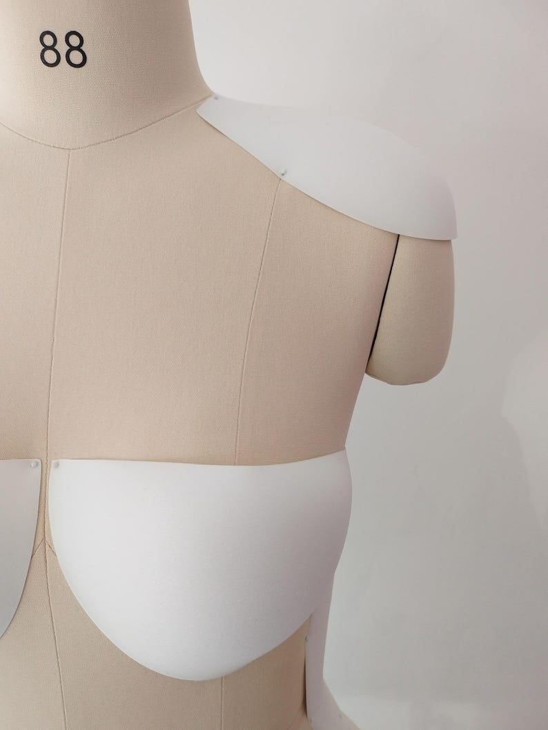 May include: A beige dress form with white foam shoulder and bust pieces attached. The dress form has the number 88 printed in black on the neck. The white foam pieces are curved and shaped to represent the form of a garment.